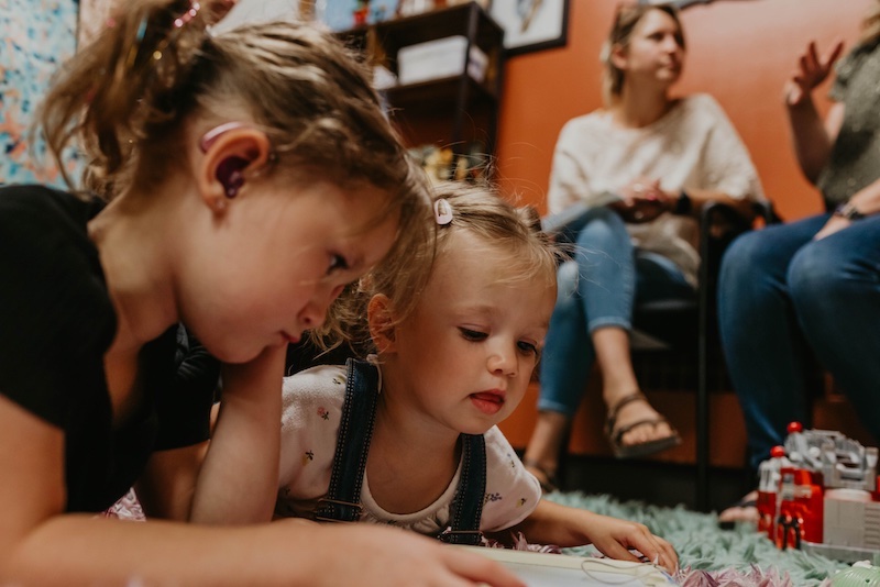 Two little girls reading a book on the floor. Two adults are sitting and taking in the background.