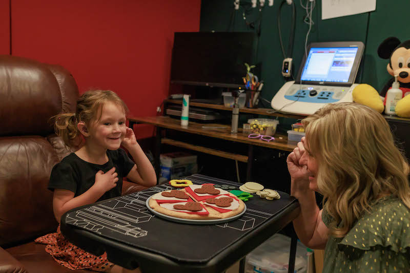 Little girl sitting with an adult and building a fake pizza together.