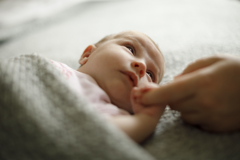 Infant laying under a tan blanket is holding someone's finger and looking up.