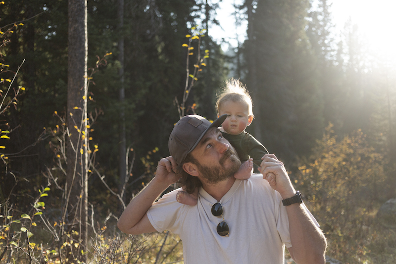 Dad and baby are walking through the woods in the sunlight. Baby is sitting on dad's shoulders.